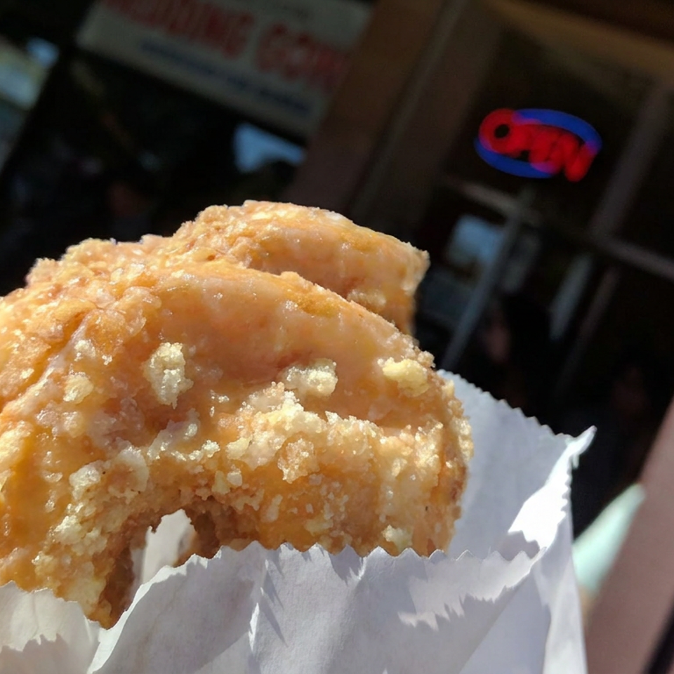 Close-up of a golden, crispy fried pastry held in paper, with a blurred restaurant interior and people in the background.